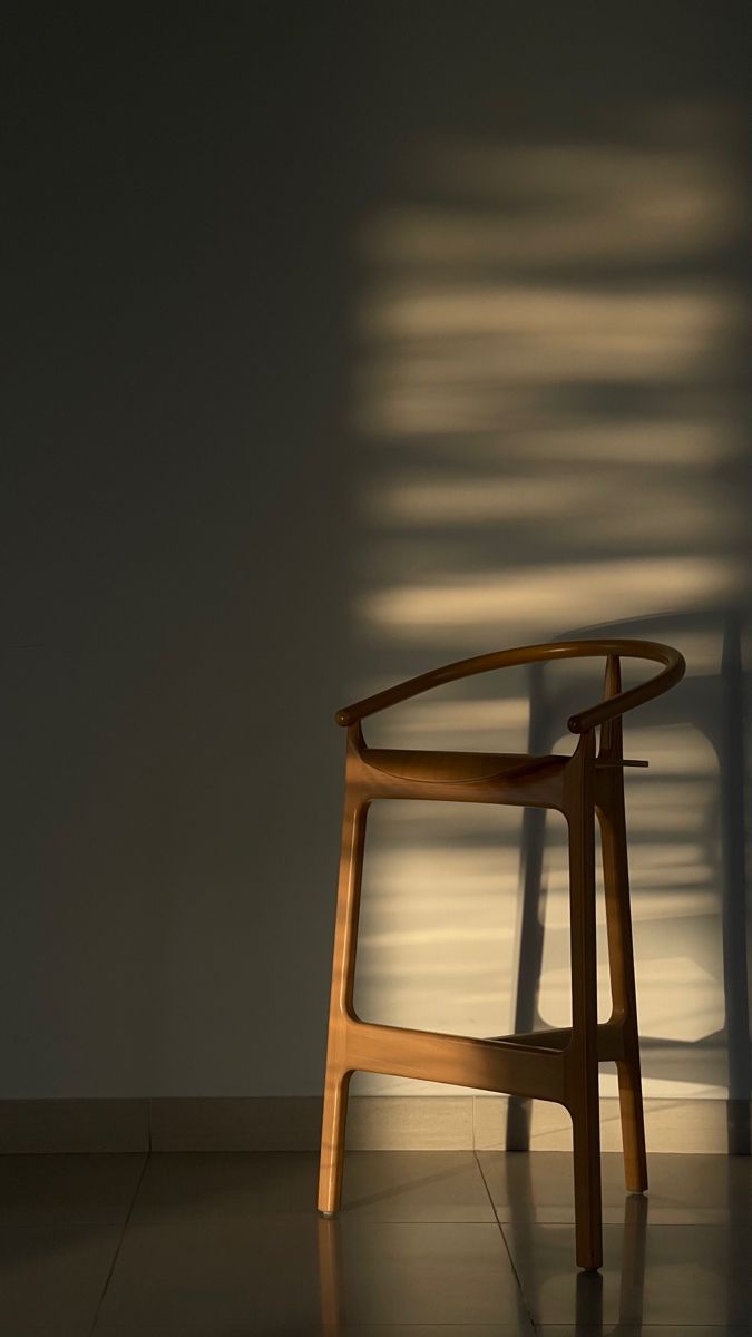 Wooden bar stool lit by soft louvred window light against a pale wall
