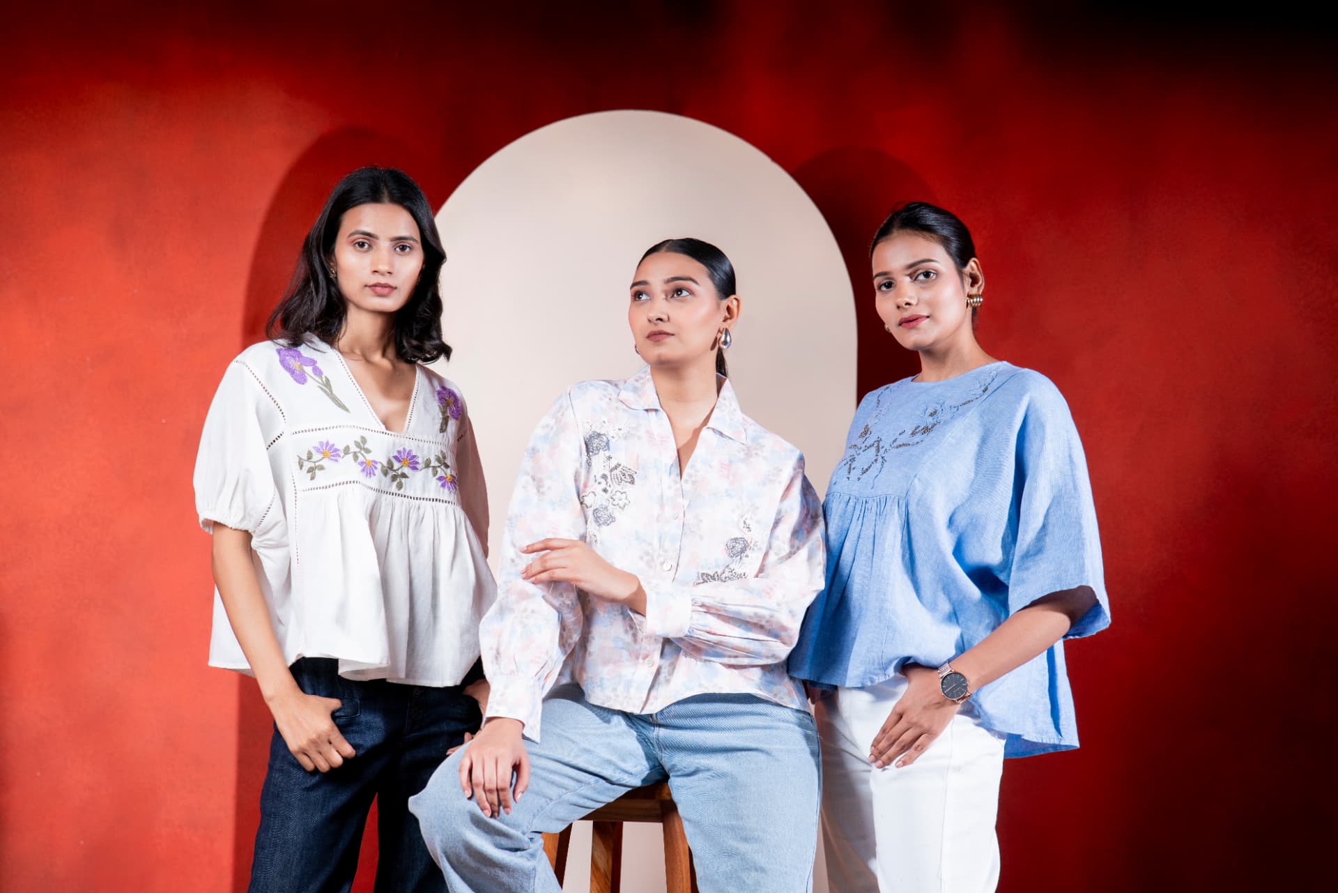 Three models in embroidered linen tops against a red studio backdrop